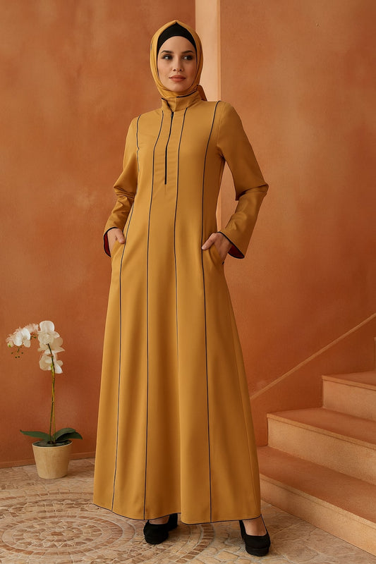 Woman wearing a mustard yellow abaya standing in a room with an orange wall and a plant.