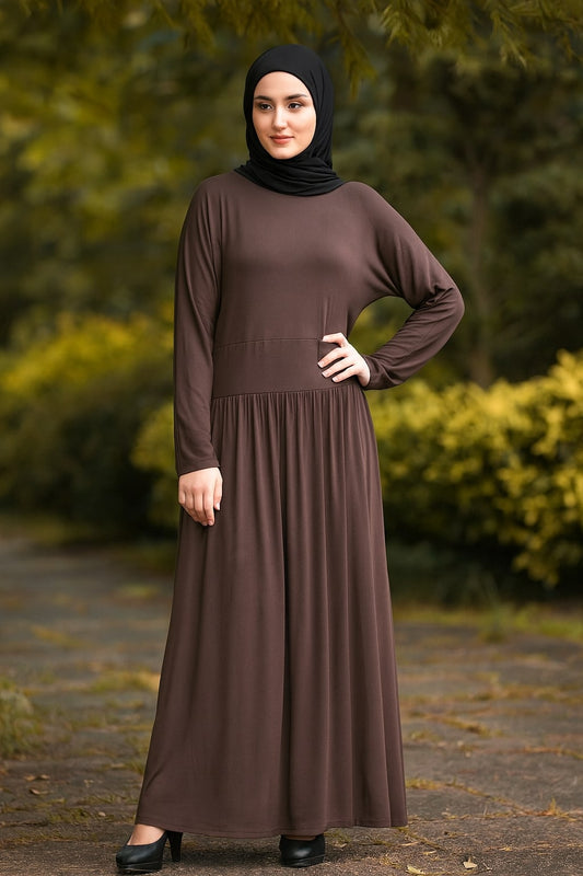 Woman wearing a brown dress and black hijab standing outdoors with greenery in the background