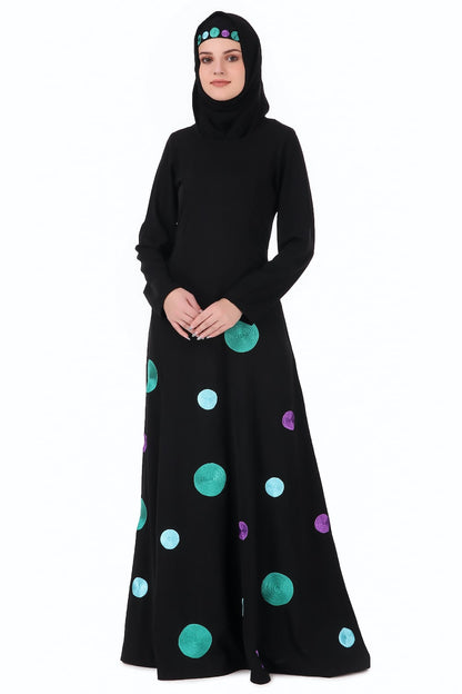 A woman wearing a black Nida abaya with round neckline and straight sleeves. The abaya features a front panel with shiny big and small polka dots embroidered below the waist line, and it has utility pockets on the sides.