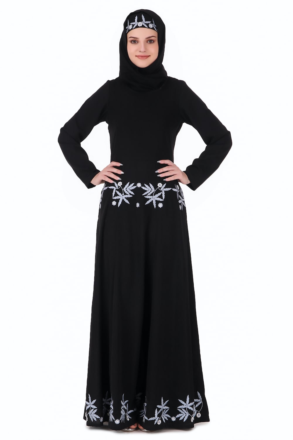 A woman wearing a black flared abaya with floral embroidery around the multi-panel bottom, paired with a matching square or rectangle hijab.