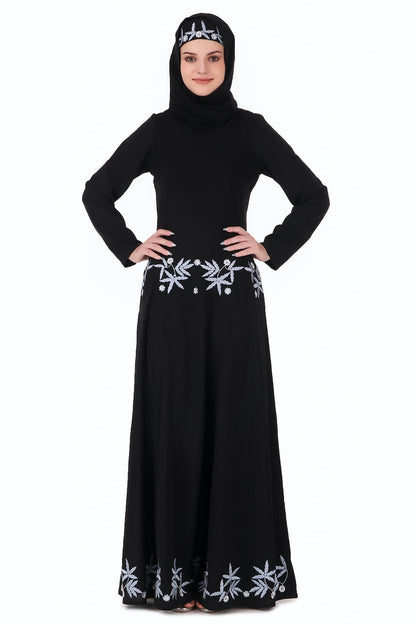 A woman wearing a black flared abaya with floral embroidery around the multi-panel bottom, paired with a matching square or rectangle hijab.