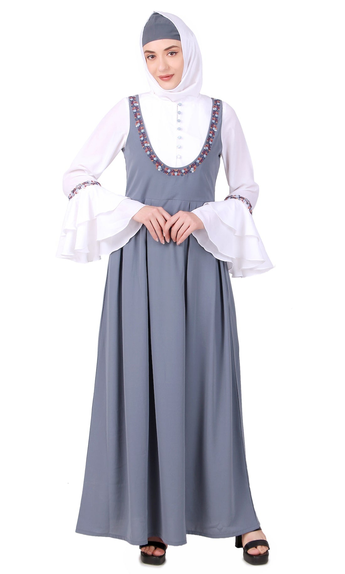 A model wearing a dual color dungaree look embroidered abaya with straight tiered cuff sleeves and floral bottom, paired with a white inner layer and a matching headscarf.