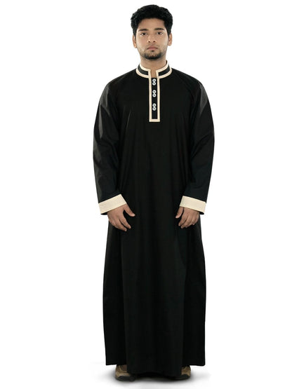 A man wearing a black galabiyya with a V-neck collar, front open placket with buttons, and utility pockets on the sides. He is also wearing cream-colored trousers.