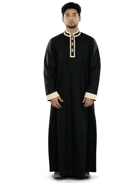 A man wearing a black galabiyya with a V-neck collar, front open placket with buttons, and utility pockets on the sides. He is also wearing cream-colored trousers.
