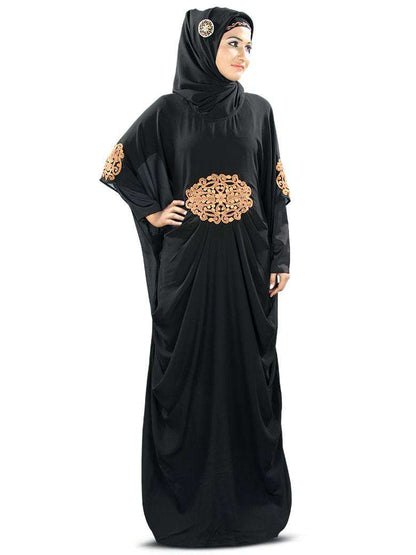 A woman wearing a black Kaftan with copper embroidery, featuring a round neckline, pleats at the waist, and matching embroidery at the sleeve ends. She is also wearing a headscarf and glasses.