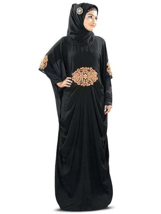 A woman wearing a black Kaftan with copper embroidery, featuring a round neckline, pleats at the waist, and matching embroidery at the sleeve ends. She is also wearing a headscarf and glasses.