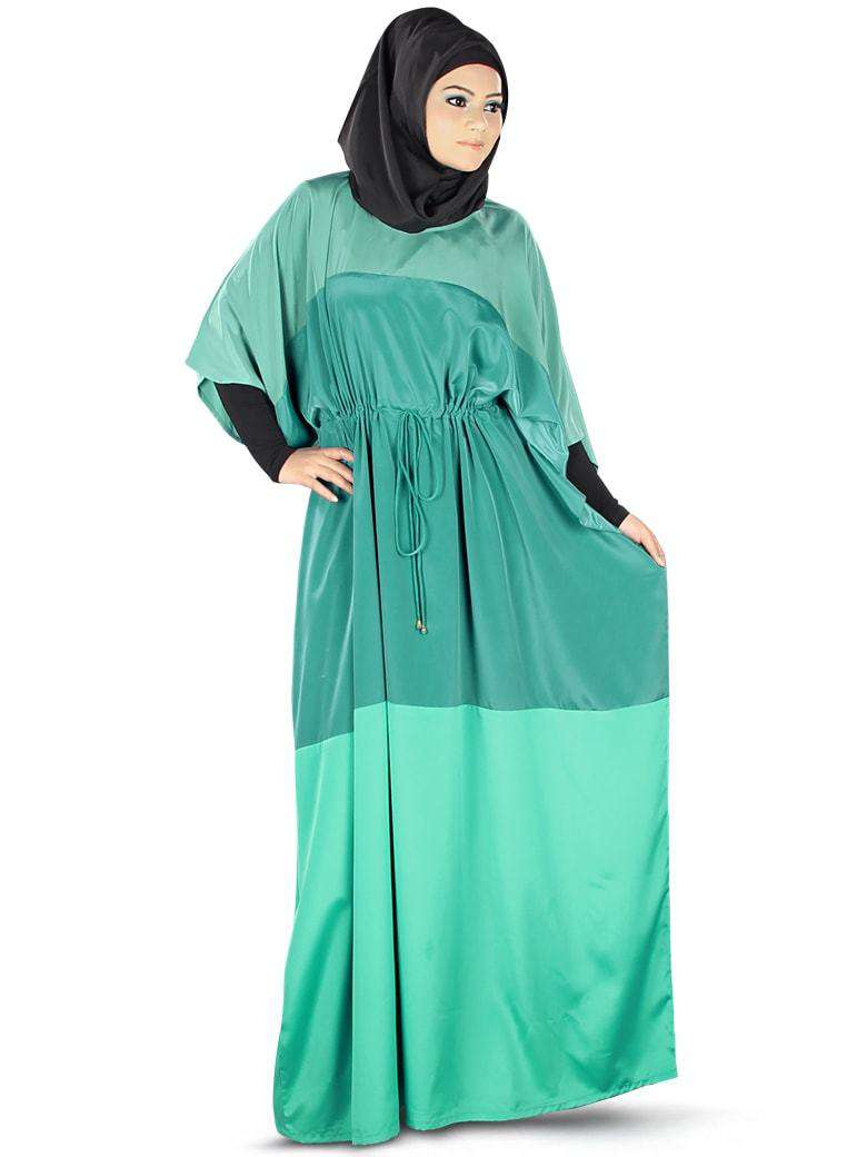 A woman wearing a green Kaftan with a round neckline, half sleeves, and an adjustable tying cord at the waist. She is also wearing a black hijab and band.
